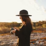 A woman with a camera stands outdoors in a sunlit, scenic landscape.