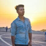 A young man in casual attire stands outdoors during a golden sunset in New Delhi, India.